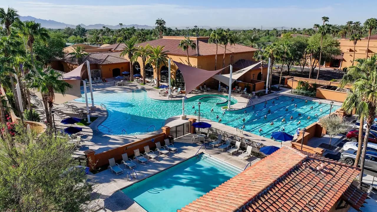 Aerial view of the PebbleCreek resort-style pool and lap lanes beside a tile-roofed clubhouse, with palm trees and mountains under a clear blue sky